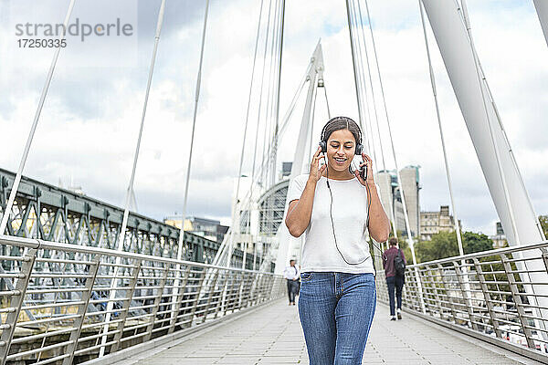 Lächelnde junge Frau  die beim Gehen auf einer Brücke über Kopfhörer Musik hört