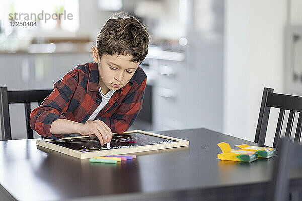 Junge zeichnet mit Kreide auf einer Schiefertafel am Tisch