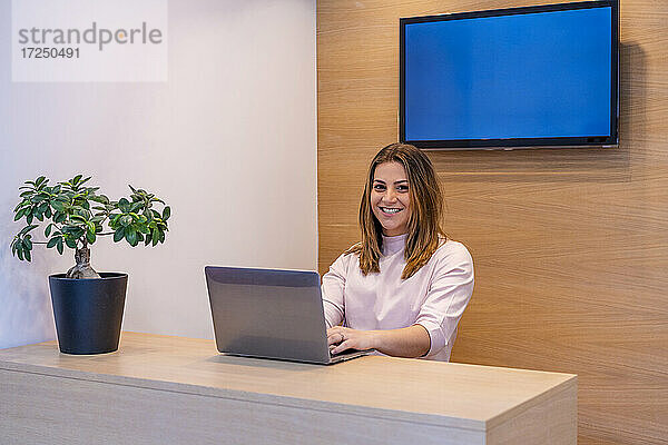 Lächelnde Empfangsdame arbeitet am Laptop am Schreibtisch in einer medizinischen Klinik