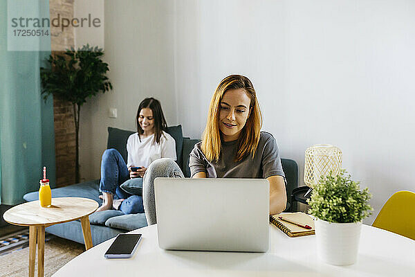 Junge Frau benutzt einen Laptop  während sie am Tisch im Wohnzimmer sitzt