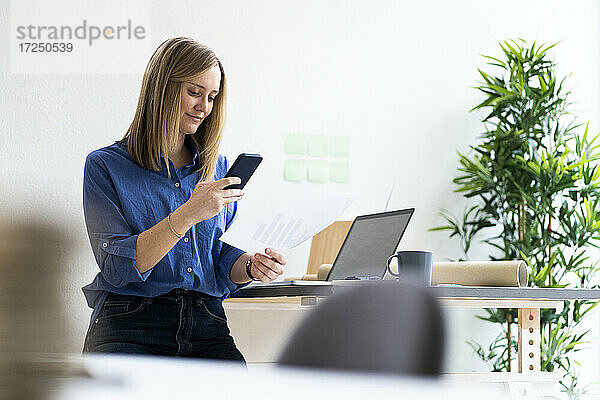 Lächelnde Geschäftsfrau  die bei der Arbeit im Büro auf ihr Mobiltelefon schaut