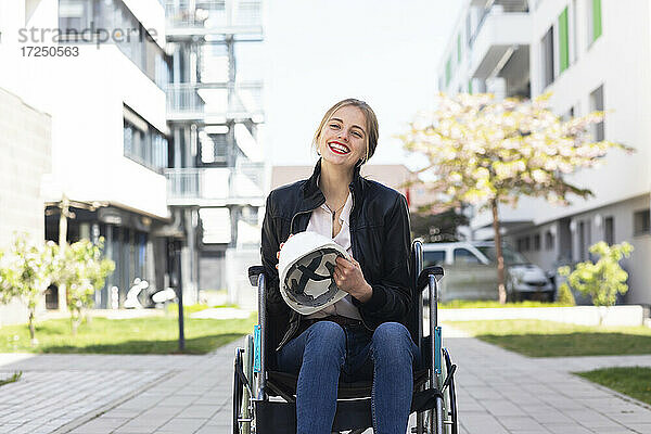 Behinderte Frau sitzt im Rollstuhl in einem Wohngebiet und hält einen Schutzhelm