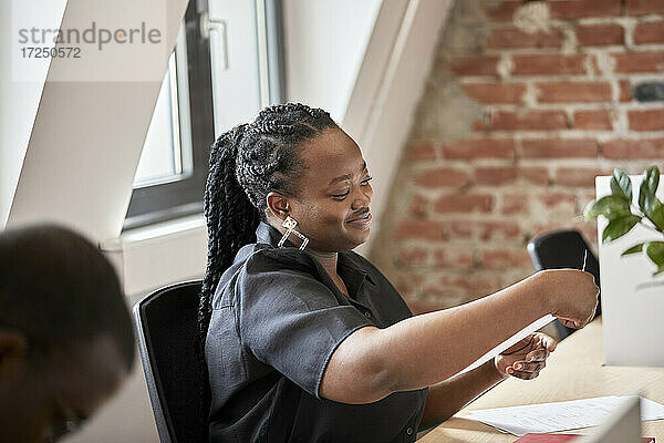 Lächelnde Geschäftsfrau mit geflochtenem Haar  die ein Dokument in einem Coworking-Büro betrachtet