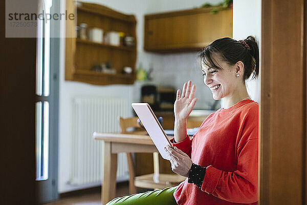 Lächelnde Frau winkt mit der Hand zu einem Videoanruf auf einem digitalen Tablet zu Hause