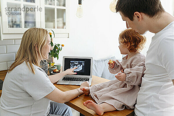Lächelnde schwangere Frau zeigt ihrer Tochter das Ultraschallbild ihres Vaters auf dem Wohnzimmertisch