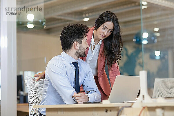 Weibliche Fachkraft schaut auf den Laptop eines männlichen Kollegen am Schreibtisch in einem Coworking-Büro