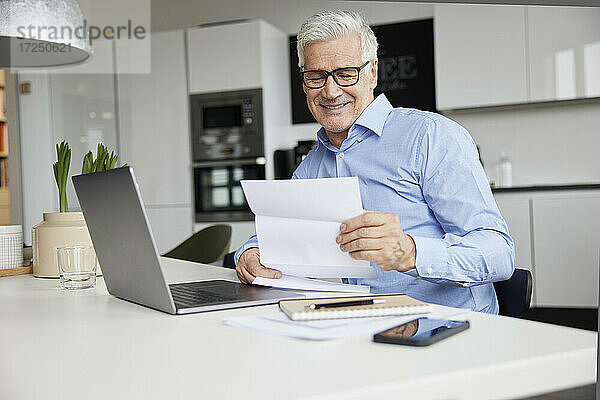 Lächelnder männlicher Unternehmer  der mit einem Laptop im Büro sitzt und ein Dokument liest