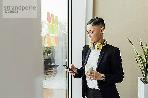 Unternehmerin hält Einweg-Kaffeebecher  während sie im Büro ihr Smartphone benutzt