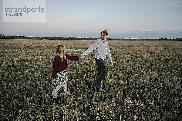 Vater und Tochter halten sich an den Händen  während sie in einem landwirtschaftlichen Feld spazieren gehen