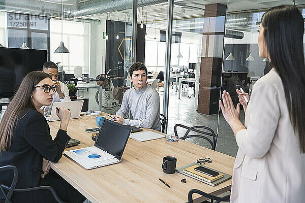 Männliche und weibliche Berufstätige schauen auf eine Geschäftsfrau  die während einer Besprechung im Büro gestikuliert