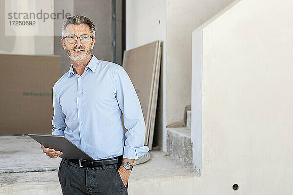 Männlicher Architekt mit Hand in der Tasche  der auf der Baustelle ein digitales Tablet hält