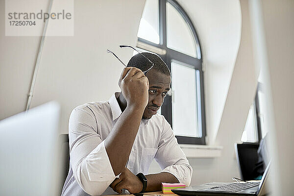Besorgter männlicher Berufstätiger  der eine Brille hält und auf einen Laptop im Büro schaut