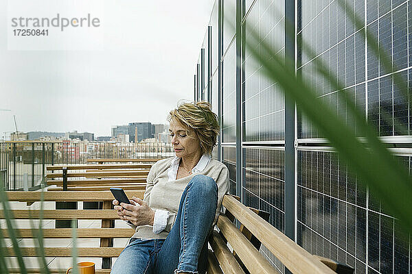 Geschäftsfrau  die ein Smartphone benutzt  während sie auf einer Bank auf einer Büroterrasse sitzt