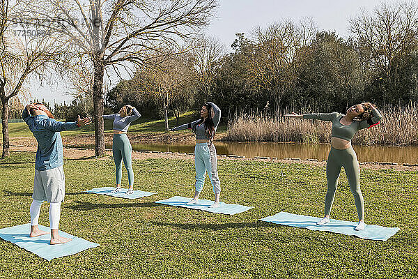 Männlicher Yogalehrer  der junge Frauen bei Dehnungsübungen auf einer Matte an einem sonnigen Tag anleitet