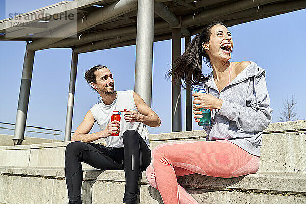 Lächelnder Mann mit Wasserflasche  der neben einer lachenden Freundin auf den Stufen eines sonnigen Tages sitzt