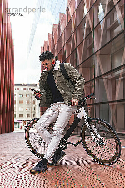 Männlicher Berufstätiger  der sein Smartphone benutzt  während er sich auf dem Gehweg an ein Fahrrad lehnt