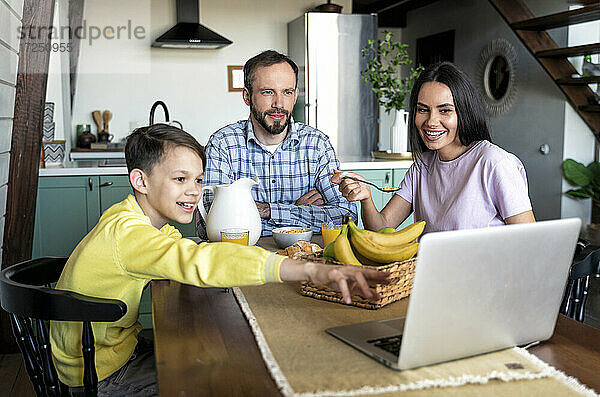 Familie schaut auf den Laptop  während sie zu Hause am Esstisch frühstücken