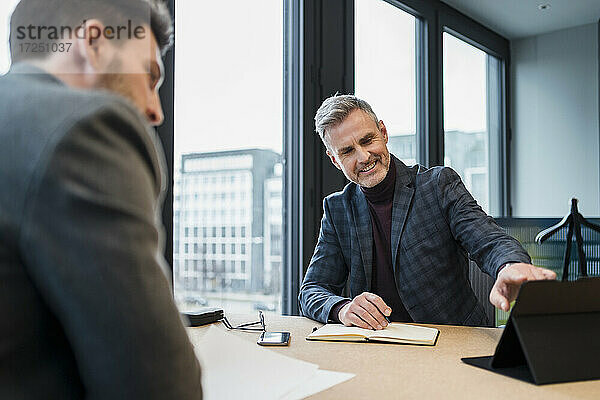 Lächelnde männliche Fachkraft  die mit einem Kollegen am Arbeitsplatz über ein digitales Tablet diskutiert