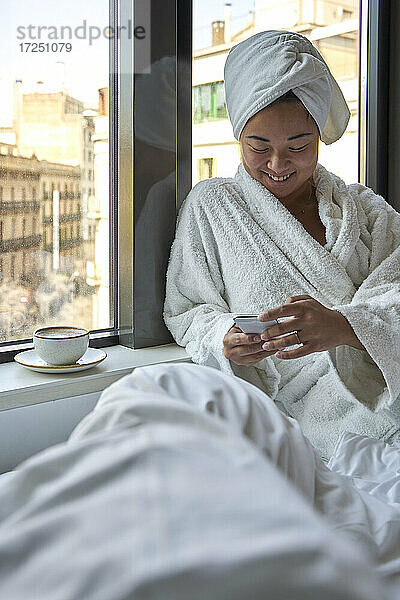 Lächelnde Frau  die ein Smartphone benutzt  während sie auf einem Hotelbett sitzt