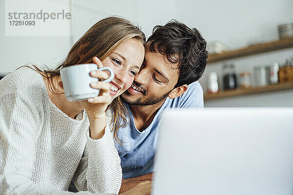 Glücklicher Mann  der seine Freundin umarmt und auf einen Laptop in der Küche schaut