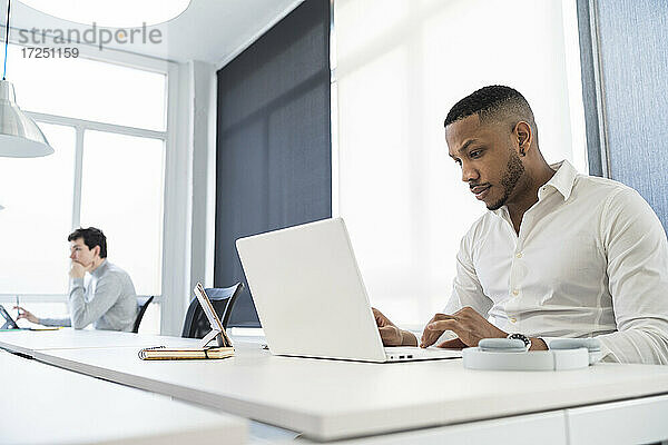 Geschäftsmann arbeitet am Laptop mit einem männlichen Kollegen im Hintergrund in einem Coworking-Büro