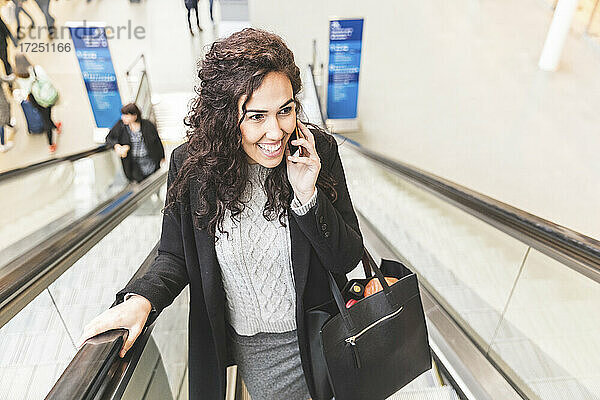 Lächelnde weibliche Fachkraft  die mit einem Mobiltelefon spricht  während sie auf einer Rolltreppe nach oben fährt