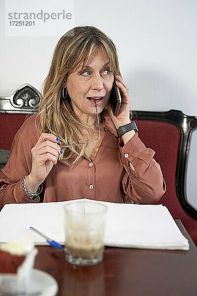 Geschäftsfrau  die wegschaut und mit einem Smartphone spricht und eine Brille in einer Cafeteria hält