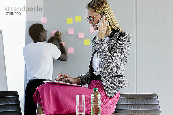 Geschäftsfrau  die mit einem Smartphone spricht  während ein männlicher Kollege im Hintergrund im Büro arbeitet