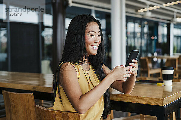 Lächelnde weibliche Fachkraft  die ein Smartphone benutzt  während sie in einer Büro-Cafeteria sitzt
