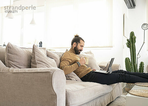Mann mit Kaffeetasse und Tablet-PC  während er zu Hause auf dem Sofa sitzt