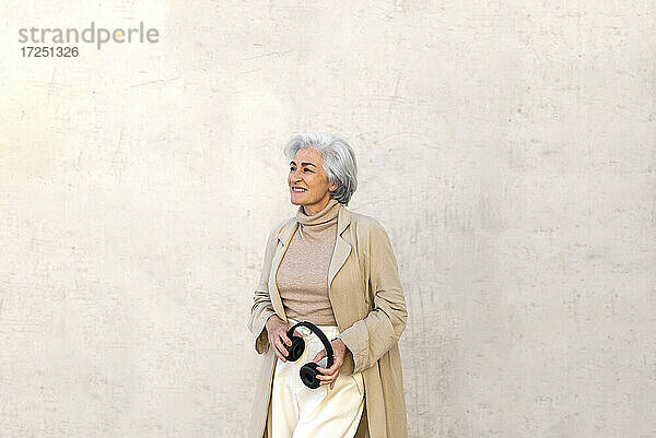 Frau mit grauem Haar träumt  während sie Kopfhörer vor einer Wand hält