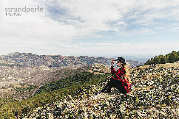 Junge Frau mit Hut  die in den Ferien auf einem Berg sitzt und nachdenkt