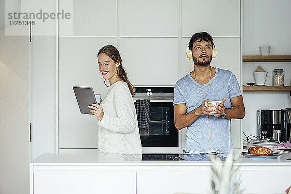 Lächelnde junge Frau  die ein Tablet benutzt  während ein Mann in der Küche Musik hört