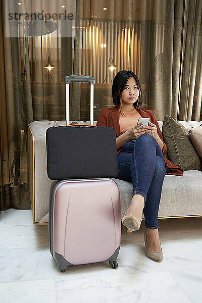 Junge Frau mit Mobiltelefon auf Sofa sitzend bei Gepäck im Hotel
