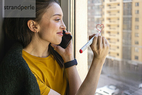 Junge Frau  die ein Herz zeichnet  während sie zu Hause mit dem Handy telefoniert