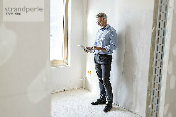 Männlicher Unternehmer  der ein digitales Tablet benutzt  während er an einer Wand auf einer Baustelle steht
