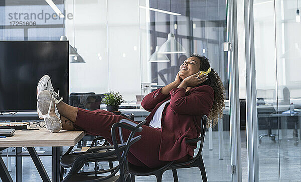 Lächelnde Geschäftsfrau hört Musik über Kopfhörer  während sie mit hochgelegten Füßen auf einem Stuhl im Büro sitzt