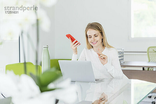 Lächelnde Geschäftsfrau mit Mobiltelefon gestikuliert während einer Videokonferenz am Laptop am Schreibtisch