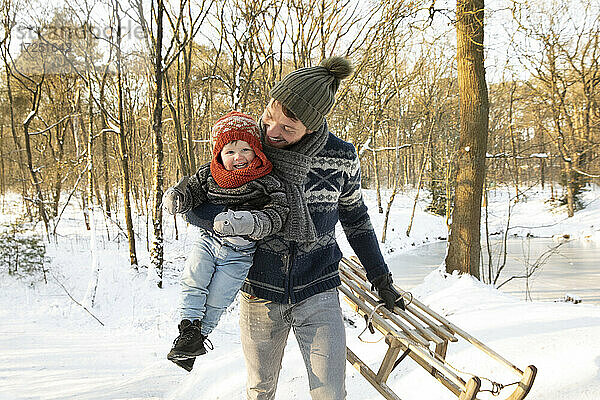 Glücklicher Vater  der seinen Sohn trägt  während er den Schlitten im Winter im Schnee hält