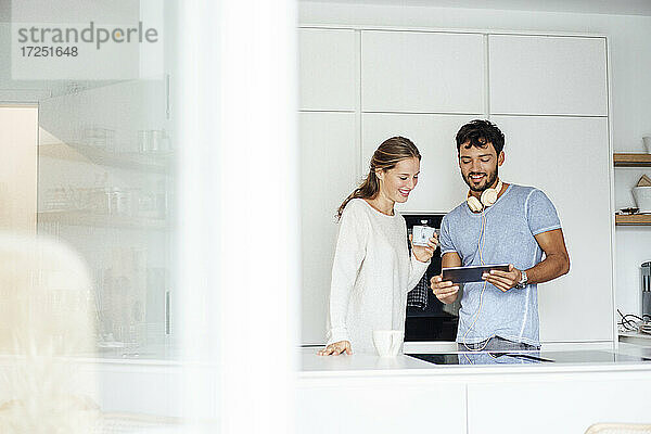 Lächelndes junges Paar  das in der Küche stehend über ein digitales Tablet diskutiert