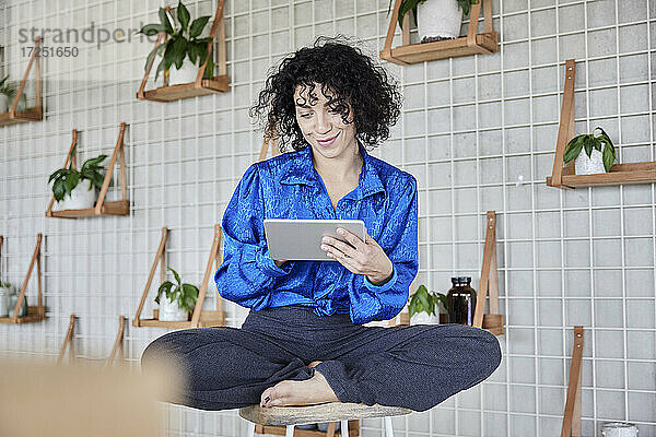 Lockenhaarige Frau sitzt auf einem Hocker und benutzt ein digitales Tablet