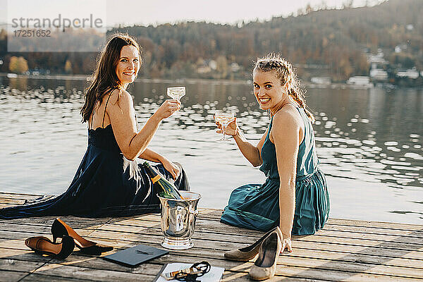Lächelnde Veranstaltungsplanerinnen sitzen mit Champagner auf einem Steg über dem See