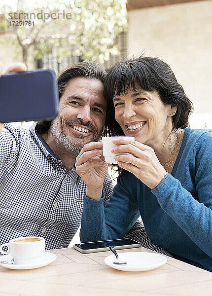 Lächelndes Paar  das ein Selfie mit dem Handy macht  während es auf der Terrasse Kaffee trinkt