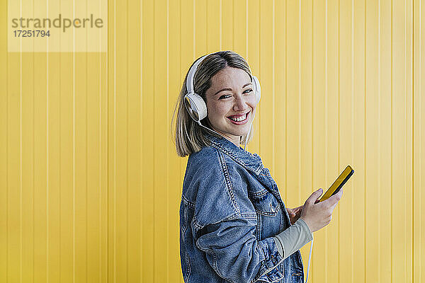 Lächelnde Frau mit Mobiltelefon hört Musik über Kopfhörer an einer gelben Wand