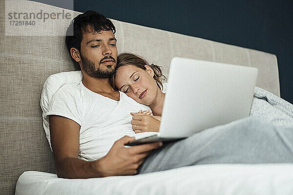 Junger Mann benutzt Laptop bei seiner Freundin  die zu Hause auf dem Bett schläft