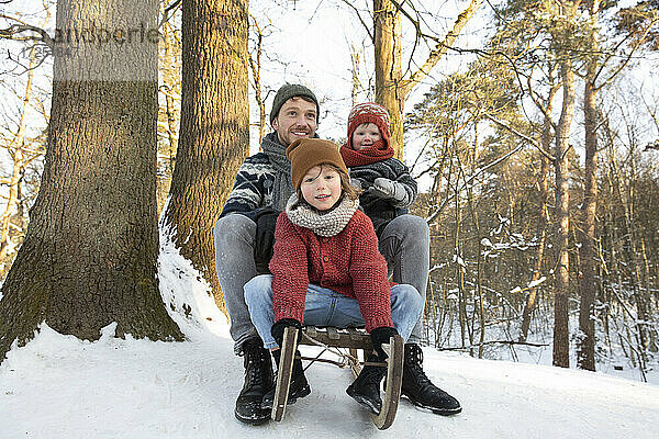 Vater auf Schlitten sitzend mit Söhnen in der Nähe des Baumstamms im Winter