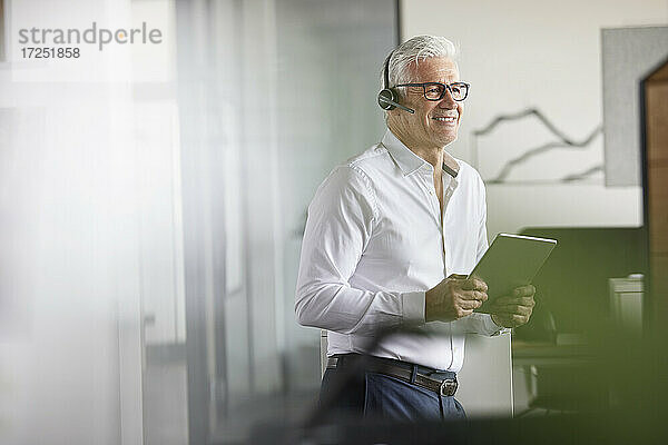 Lächelnder männlicher Fachmann mit drahtlosem Headset und digitalem Tablet  der im Büro wegschaut