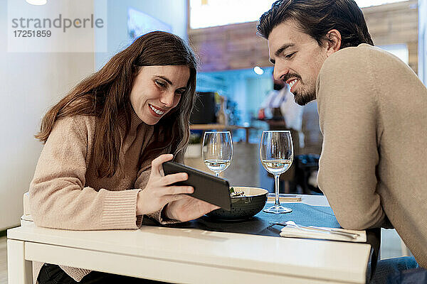 Lächelnde junge Frau zeigt ihrem Freund im Restaurant ihr Smartphone