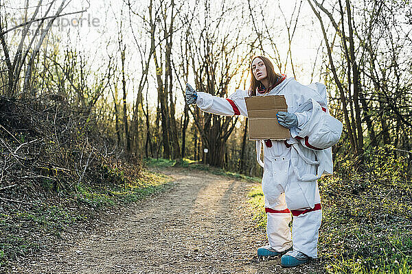 Weibliche Astronautin im Raumanzug gestikuliert  während sie einen Karton auf einem Feldweg hält