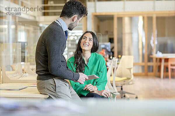 Lächelnde Geschäftsfrau  die einen männlichen Kollegen ansieht  der sich im Büro auf den Schreibtisch stützt